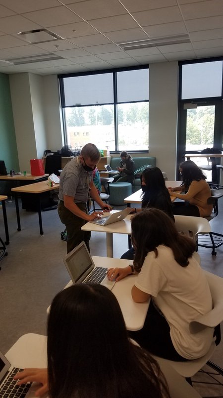 A teacher in a classroom of students looks at one student's laptop